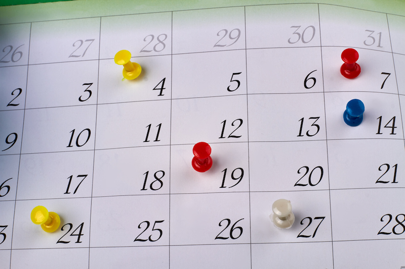 Many colorful push pins on a monthly paper calendar page. Deadlines and events planning concept.