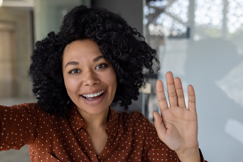 Beautiful business woman with curly hair wearing looking at smartphone camera, waving her hand in greeting gesture, talking with friends and colleagues, using video call app