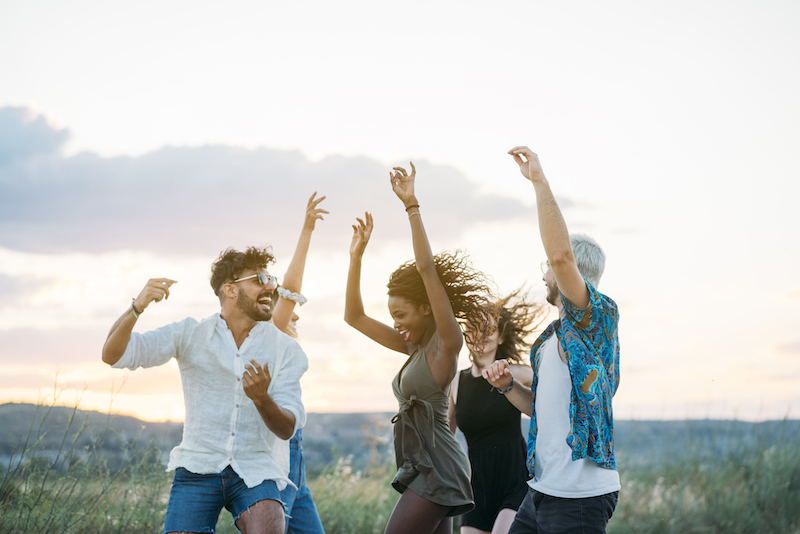 Group of young people in casual outfits laughing and dancing while having fun in beautiful countryside together