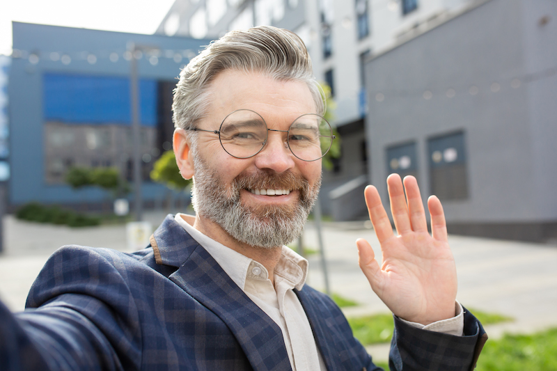 Experienced senior businessman in a sharp suit waving hello outside an office, embodying professionalism and friendliness.