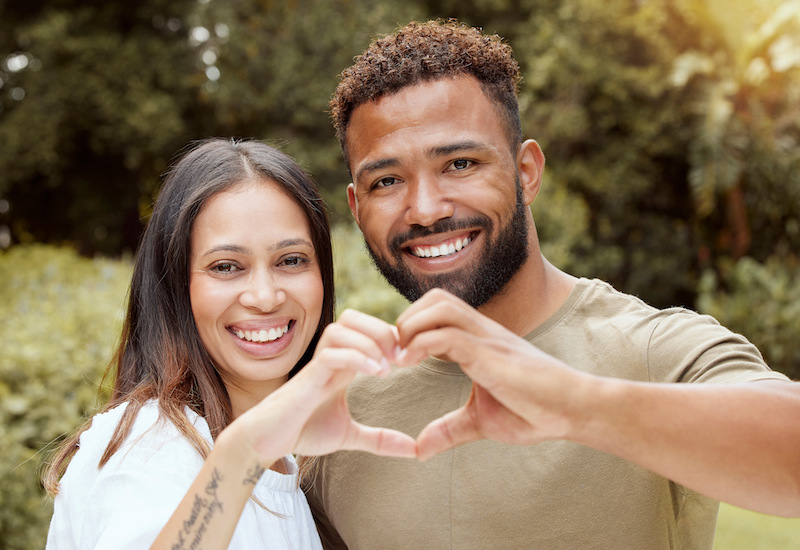 Couple, happy and hand heart sign in a nature park showing love and a smile outdoor.