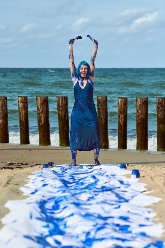 Young woman performance artist in dark blue dress smeared with blue gouache paints happily dancing hands up on beach. Body painting and body art. Canvas for expressive painting with indigo gouache