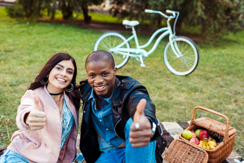 Portrait of happy interracial couple showing thumbs up while sitting on a grass in the park
