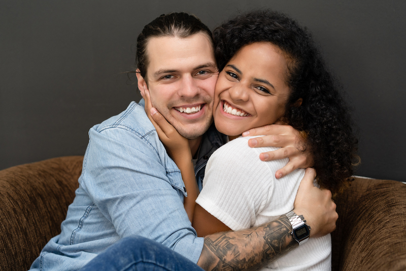 Smiling mixed-race young couple hugging and looking at camera on the chair at home. Relationship concept with boyfriend and girlfriend together