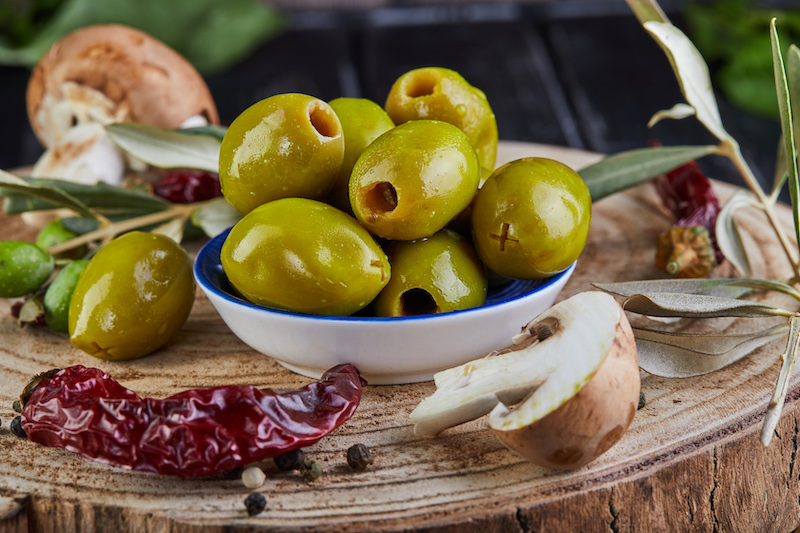 Still life of green fresh olives, red pepper and fresh mushrooms with olive tree leaves on a dark wooden background close up