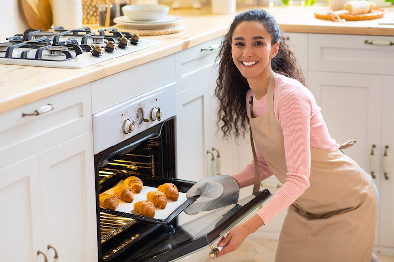 Happy Young Woman Taking Out Tray With Fresh Baked Croissants From Oven In Kitchen