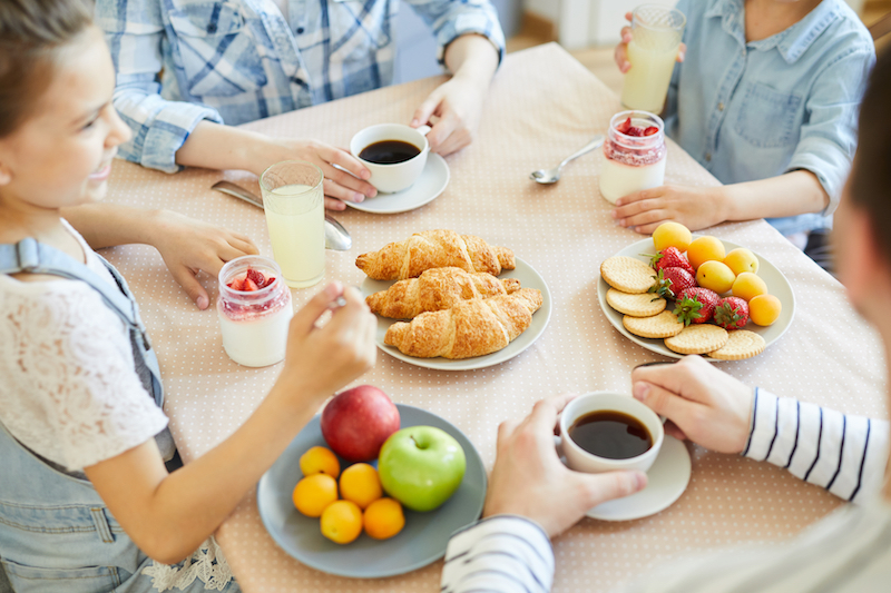 Parents having coffee and little siblings eating strawberry dessert by table served with fruits, croissants and biscuits