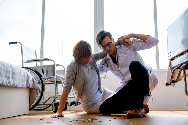 Doctor man help to support senior woman who fall from wheelchair to floor of patient room and medicine are spread on the floor.