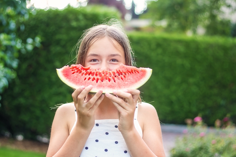 Happy playful preteen girl holding big slice of watermelon like smile in front of her face in garden outdoors. Summer lifestyle.