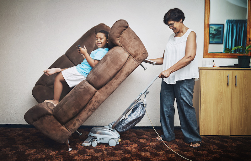 Shot of a grandmother vacuuming under a couch which her grandson is lying on at home.