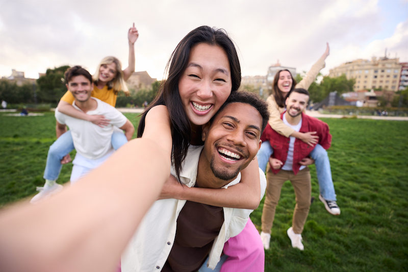 Multirracial group of friends taking selfie picture walking the park hugging by couples and smiling. Happy young people smiling together at the camera outside. University students having fun in college campus. Youth culture.