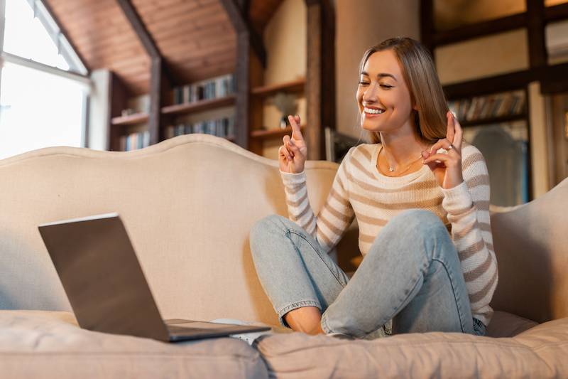 Making Wish. Portrait of smiling hopeful woman crossing fingers, anticipating something with faithful expressions using pc sitting on sofa, excited lady pleading for good luck and positive result