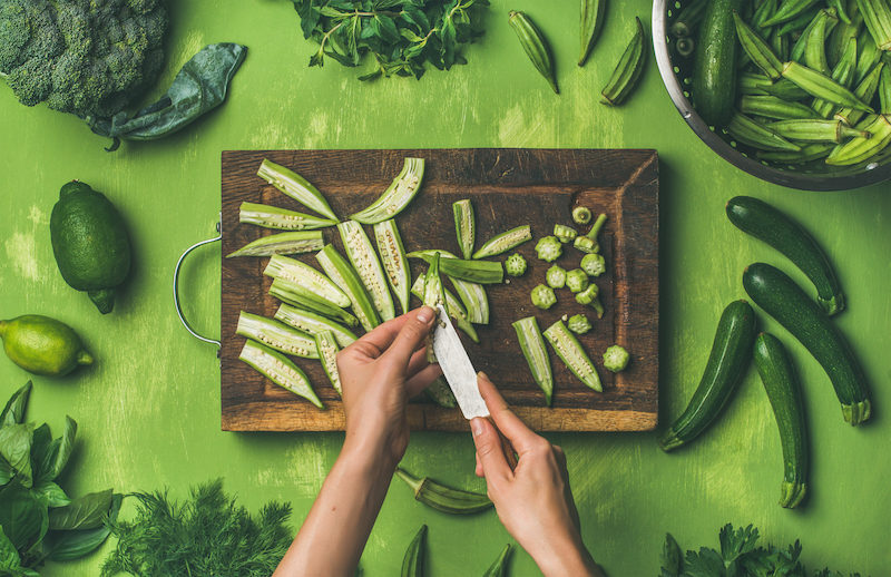 Healthy vegan cooking ingredients. Flay-lay of woman hands cutting green vegetables and greens over wooden green background, top view. Clean eating, vegetarian, detox, dieting food concept