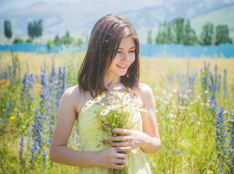 Beautiful woman in yellow dress standing on blooming field with flowers and holding bouquet of chamomile and smiling