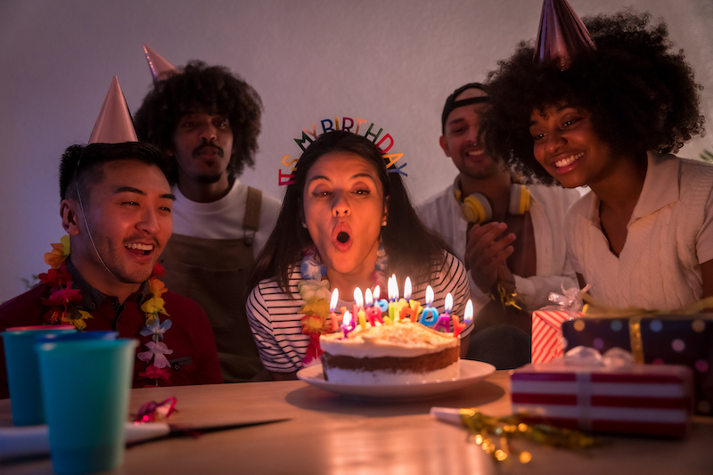 Multi-ethnic group of friends at a birthday party on the sofa at home with a cake and gifts, blowing out the candles with the lights off