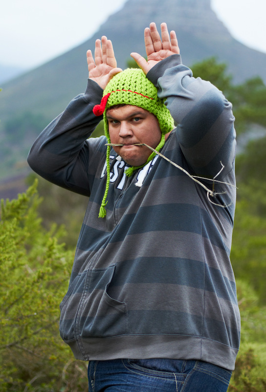 An overweight man posing with his hands in bunny ears and a twig in his mouth while wearing a playful cap