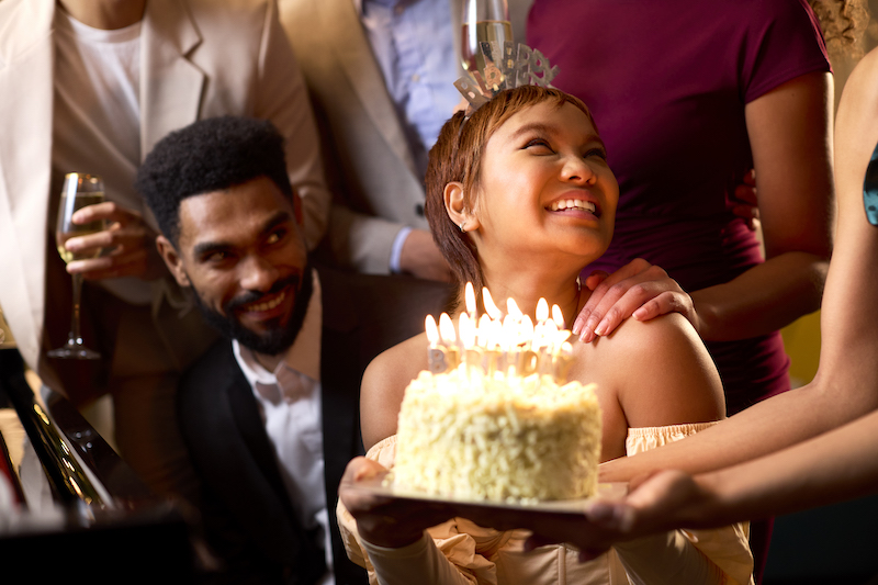 Close Up Of Group Of Friends Around Piano Celebrating Woman's Birthday With Cake