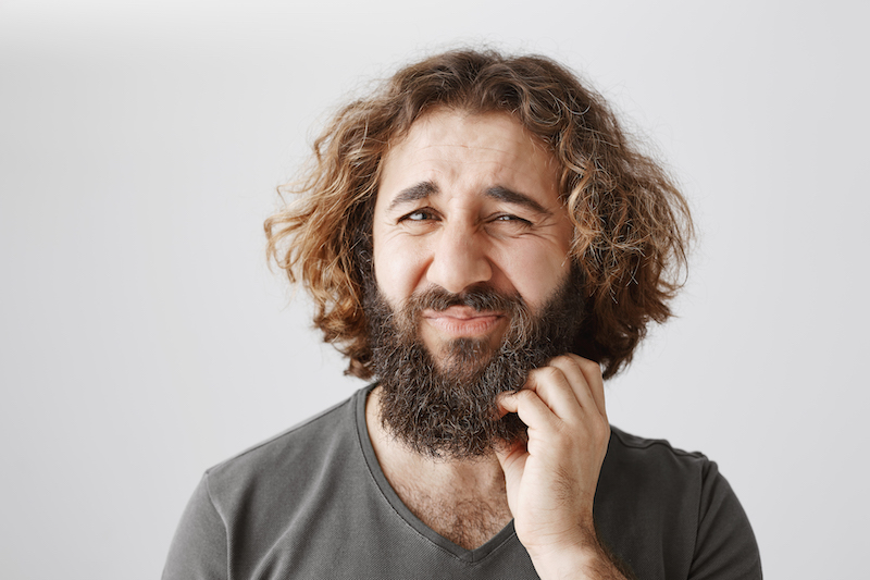 Indoor portrait of hesitating uncertain eastern male with curly hair frowning and scratching beard, expressing indifference and dislike, being uninterested in offer and wanting to reject it. Copy space