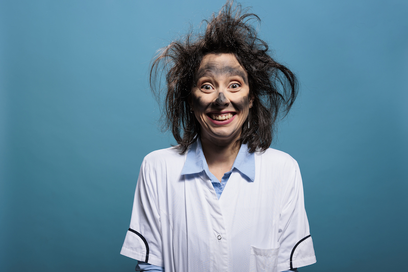 Portrait of crazy chemist with dirty face and messy hair grinning dreadful at camera while on blue background. Mad scientist with dreadful smile and stupid look after chemical laboratory explosion.