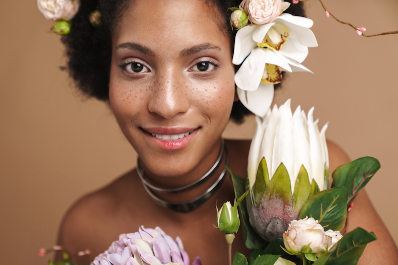 Portrait of young half-naked freckled african american woman posing with flowers isolated over beige background