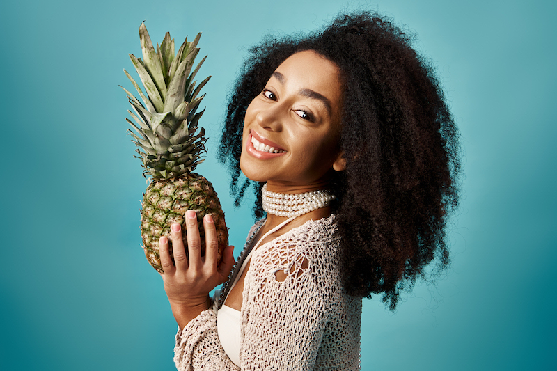 Young African American woman in swimsuit holding pineapple against blue backdrop.