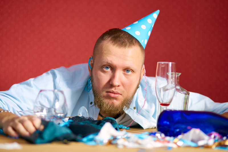 Tired man at table with blue cap in messy room after birthday party, tired man after party at home