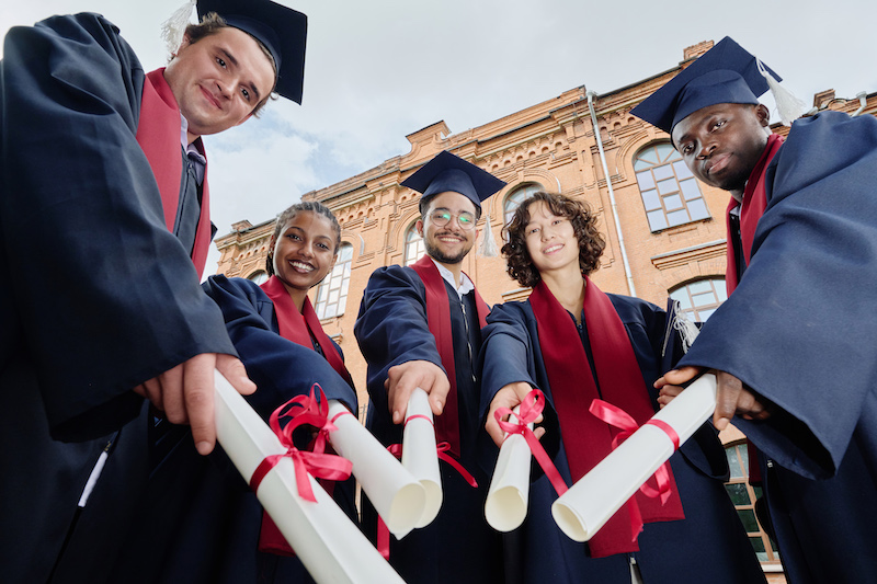 Low angle view of group of graduated students with diplomas smiling at camera while standing outdoors