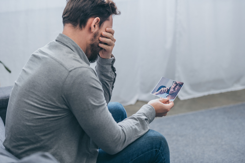 man sitting and crying while holding a male portrait at home, grieving disorder concept