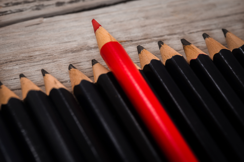 Red pencil stands out from the crowd of black pencil on a wooden white background.