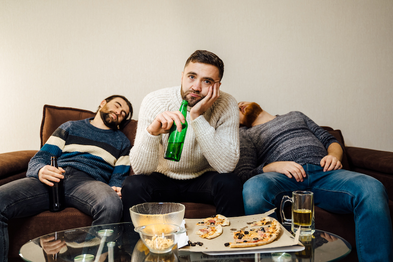 Young bored man drinking beer from bottle siting on couch watching using TV. Looking disinterested and sleepless. while his friends slept beside him