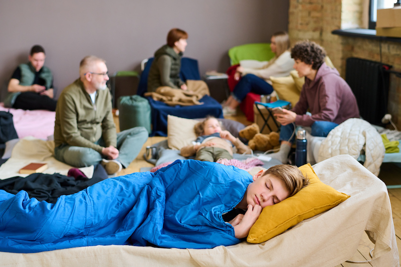 Adolescent boy keeping his head on pillow while sleeping under blue blanket against group of people talking on their beds