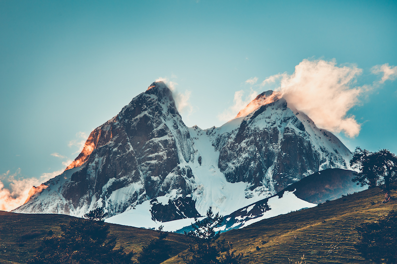 Sunset over the snowy mountain peaks with misty cloud and green hill.