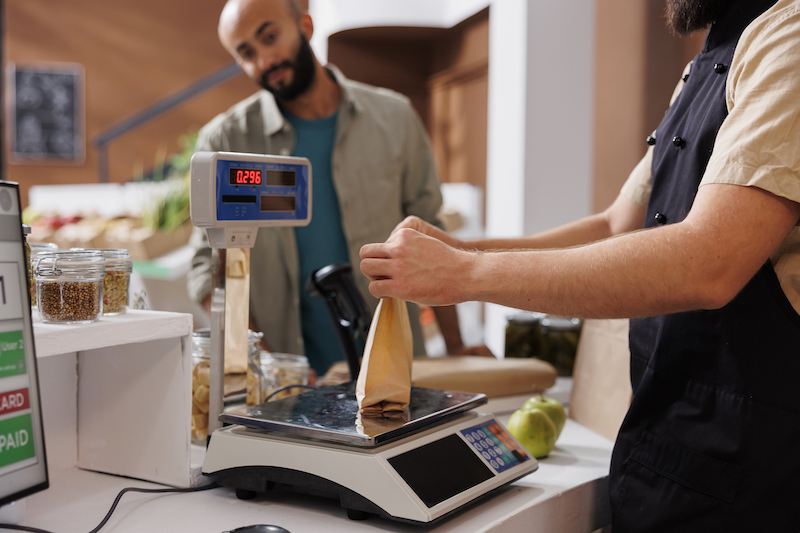 At checkout counter vendor places brown paper bag on weighing scale to check value for customer. 